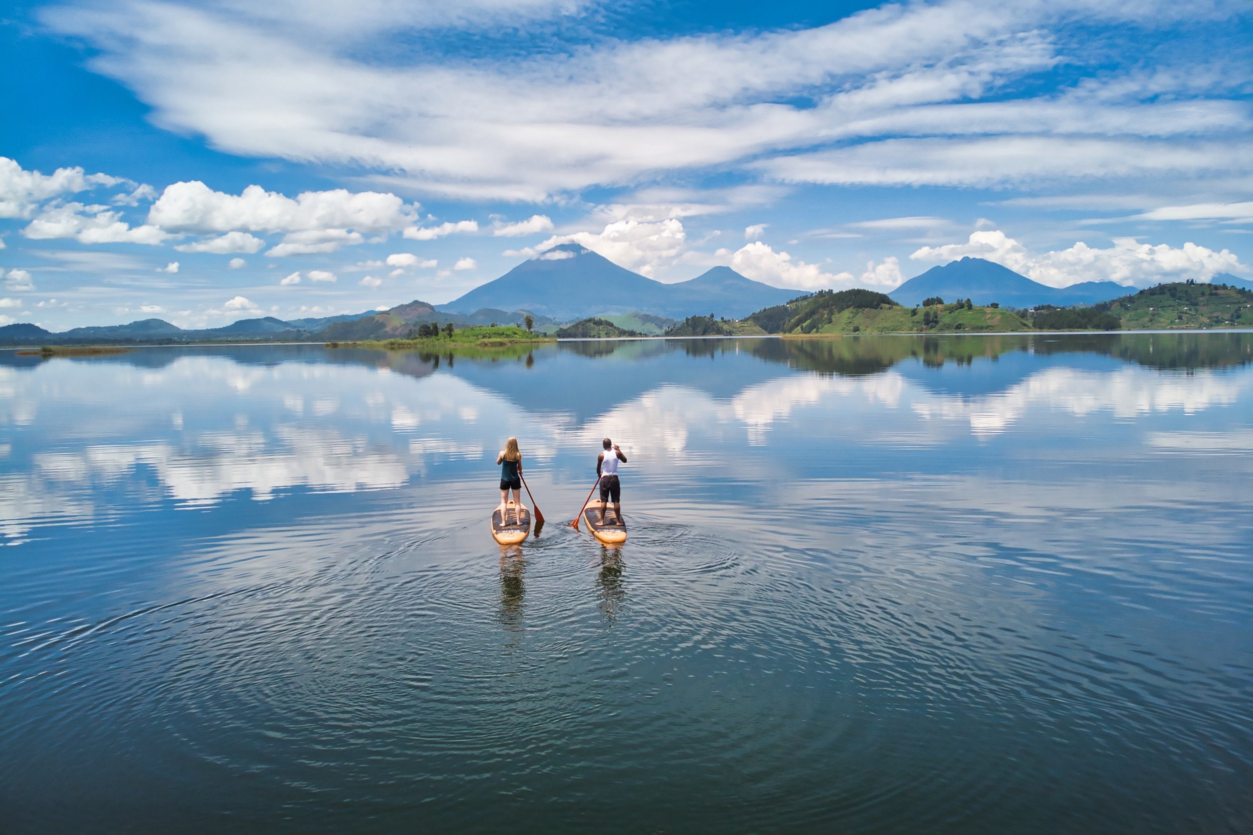 Two tourists paddleboarding on Lake Mutanda, Uganda, with the dramatic peaks of the Virunga Mountains reflecting in the calm water.