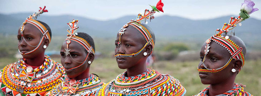 Four members of a Ugandan tribe wearing traditional colorful beaded jewelry and headdresses, side-by-side.